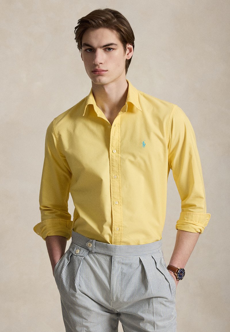 Jeune homme aux cheveux bruns portant une chemise jaune à manches retroussées, un pantalon rayé taille haute et une montre marron, posant sur un fond neutre.