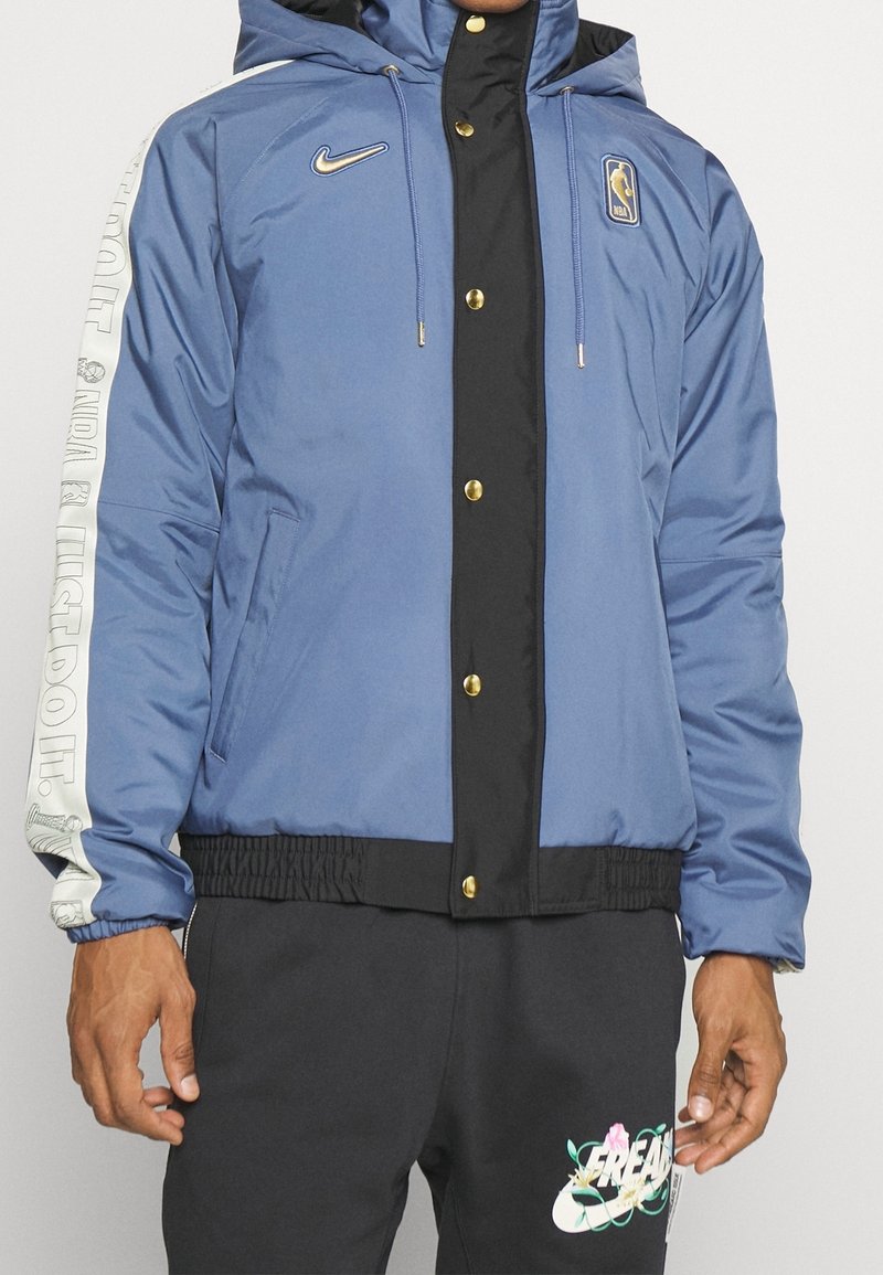 Veste à capuche Nike NBA bleue et noire avec des boutons dorés et un texte blanc "NBA & Just Do It." sur la manche, portée avec un pantalon noir avec un logo floral Nike.