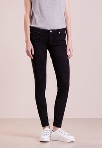 Jean skinny en velours noir avec une coupe slim et un design à cinq poches, accompagnés de baskets blanches à lacets et d'un haut gris sans manches.