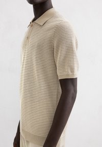 Beige knit polo shirt with a textured pattern, short sleeves, and a classic collar. Features two buttons at the neck and ribbed hem.