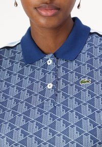 Polo bleu avec un motif géométrique en bleu clair. Il dispose d'un col, de trois boutons et d'un petit logo crocodile vert.
