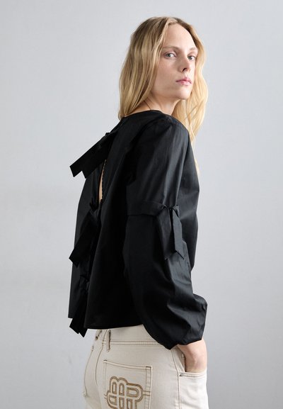 Čierna blúzka s dlhým rukávom a balónovými rukávmi, so šnúrkami na zadnej strane. Materiál pôsobí textúrovane. Spárovaná s béžovými nohavicami s výšivkou.