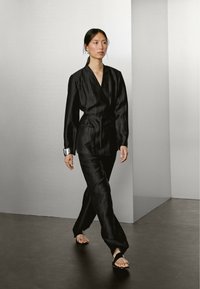 Outfit in tessuto nero strutturato con giacca avvolgente e pantaloni a gamba larga. Presenta una cintura, dettagli in argento e calzature con punta aperta.