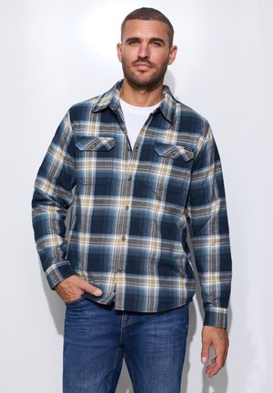 Mann trägt ein blau, grau und beige kariertes Hemd über einem weißen T-Shirt und blauen Jeans, steht mit einer Hand in der Tasche.