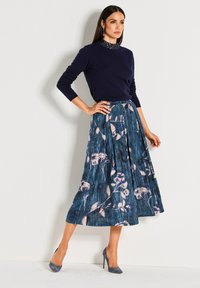 Marineblauwe sweater met een getextureerde halslijn, gecombineerd met een geplooide, bloemenprint rok in blauwe tinten. Hakken maken de outfit compleet.