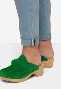 Zuecos de ante verde con un diseño redondeado, plataforma de madera clara y detalles de costura visibles. Los puños de mezclilla enrollados muestran los tobillos.