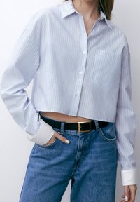 Donna che indossa una camicia a righe azzurro chiaro, cropped, con polsini bianchi, infilata in jeans blu a vita alta con una cintura nera.
