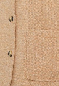 Υφασμα με υφή σε ανοιχτό πορτοκαλί χρώμα με ένα κουμπί και μια τσέπη, με πυκνό υφαντό μοτίβο που προσδίδει διακριτικό βάθος και ενδιαφέρον.