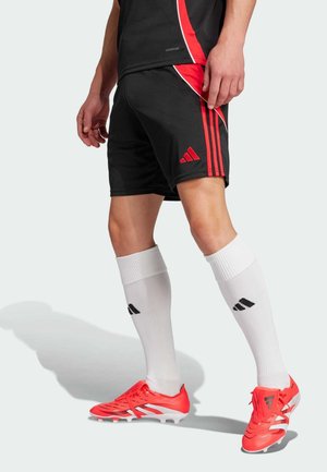 Mężczyzna sportowiec ubrany w czarne i czerwone spodenki piłkarskie, białe, sięgające kolan skarpetki oraz czerwone korki, stojący na jasnym tle.