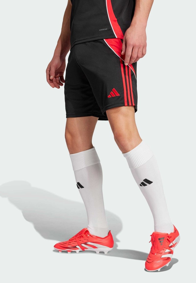 Athlète masculin portant un short de football noir et rouge, des chaussettes blanches montantes et des chaussures à crampons rouges, debout sur un fond clair.