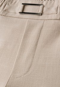 Béžová látka s jemnou texturou, se zapuštěnou přední kapsou a kovovým obdélníkovým přezkou na pásku.