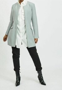 Cappotto grigio chiaro su misura con chiusura a scatto, dotato di due tasche frontali, indossato sopra una camicia bianca. Abbinato a pantaloni neri e stivali neri.