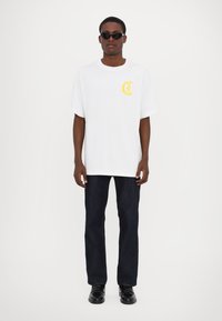 Hvit bomull T-skjorte med gult logo på brystet, avslappet passform. Kombinert med mørkeblå jeans og svarte sko. Modellen bærer svarte solbriller.