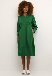Zelené šaty s výstřihem do V, tříčtvrtinovými nadýchanými rukávy a bočními kapsami. Hladká textura a délka po kolena. Nosit s bílými sandály.