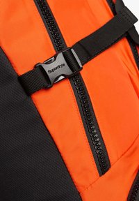 Primer plano de una mochila naranja y negra que muestra una hebilla de plástico negra con el texto "Superdry" y una cremallera negra.