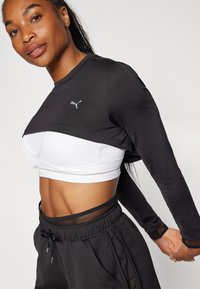 Schwarzes Langarmoberteil mit einem cropped Design über einem weißen Sport-BH, das aus einem strukturierten Stoff besteht und Mesh-Akzente an den Bündchen und am Bund aufweist.