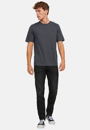 Jeune homme regardant vers la gauche, portant un t-shirt gris foncé, un jean noir et des baskets noir et blanc, debout devant un fond blanc.