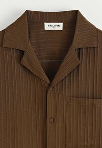 Chemise brune à côtes avec un large col et une poche poitrine, présentant une petite étiquette de marque blanche à l'intérieur du col.