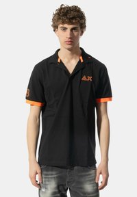 Polo nero con accenti arancioni su maniche e colletto, con dettaglio del logo e numero "8" sulla manica sinistra. Materiale in cotone morbido.