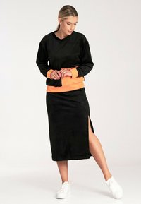 Czarna dresowa bluza z akcentami w kolorze pomarańczowym, w zestawieniu z dopasowaną czarną spódnicą z bocznym rozcięciem i białymi sneakersami.