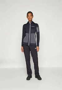 Svart jakke med glidelås, grå detaljer og flere lommer, kombinert med svarte bukser og mørke sko, designet for aktiv bruk.