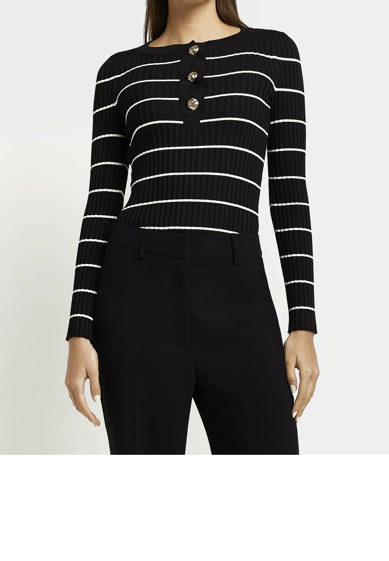 Femme portant un haut noir à manches longues côtelé avec des rayures horizontales blanches et trois boutons décoratifs dorés, associé à un pantalon noir sur mesure.
