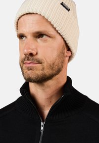 Gorro de punto color crema con textura acanalada, combinado con una camiseta negra de cuello con cremallera que presenta un collar texturizado y detalle en cremallera plateado.