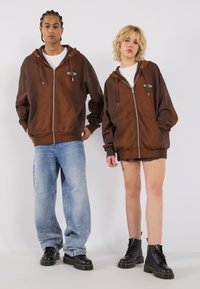Brown zip-up hoodies with drawstrings, front pockets, paired with light blue jeans and black lace-up boots. Model poses with casual stance.