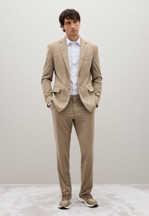 Beige Anzugjacke mit passender Hose, weißem Hemd und tanfarbenen Sneakern. Glatter Stoff, taillierte Passform, minimalistisches Design, ohne Muster oder Akzente.