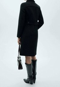 Abrigo de lana negro con diseño ajustado, cinturón abotonado en la parte trasera, combinado con botas negras hasta la rodilla y un bolso oscuro.