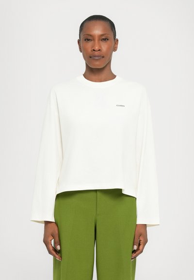 Haut blanc à manches longues avec un col rond, orné d'un petit logo "CLOSED" sur la poitrine, associé à un pantalon vert.