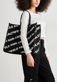 Svart tote-väska med vit "KARL LAGERFELD" logotyp, slät tyg, strukturerad form och två svarta handtag, hålls av en person.