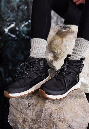 Personne portant des bottes d'hiver isolées noires avec des semelles blanches et des chaussettes en tricot grises, assise sur un grand rocher à l'extérieur.