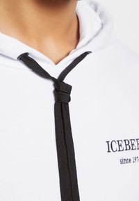 Vue partielle d'une personne portant un sweat à capuche blanc avec des cordons noirs noués et le texte "ICEBERG since 1973" sur la poitrine.