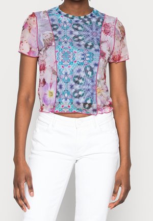 Kvinde iført en semigennemsigtig kortærmet top med blå og lyserøde blomsterprint, kombineret med hvide bukser med høj talje.