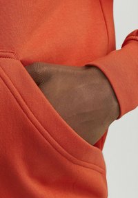 Sudadera naranja de tela suave con un bolsillo tipo canguro; una mano está colocada dentro del bolsillo, destacando el diseño de la prenda.