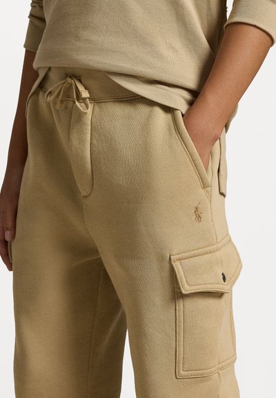 Calças de treino bege com cintura ajustável por cordão, bolsos laterais e um bolso de carga com aba. Textura suave e tecido respirável.