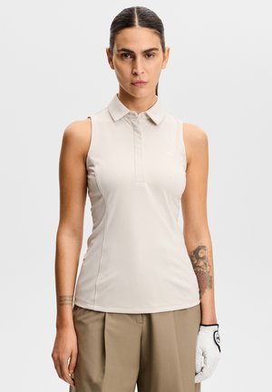 Femme aux cheveux tirés en arrière portant un polo beige sans manches, un pantalon kaki, un gant blanc et un tatouage sur son avant-bras gauche.