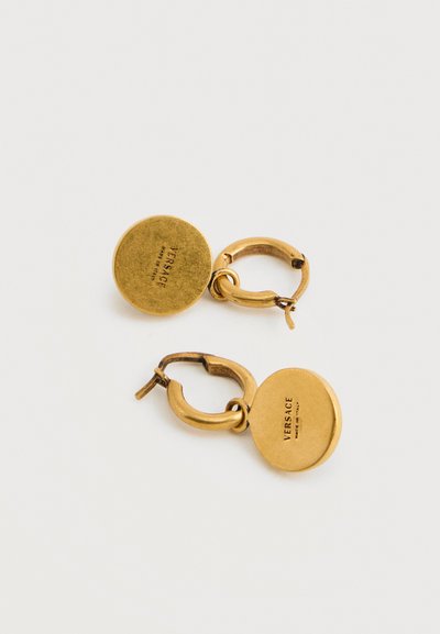 Σκουλαρίκια κρίκοι Versace σε χρυσαφί τόνο με στρογγυλά μενταγιόν με χαραγμένη την επιγραφή "VERSACE MADE IN ITALY" σε απλό λευκό φόντο.
