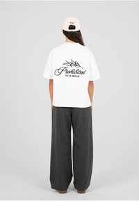 Hvit T-skjorte med svart kursiv tekst "Prohibited 10119 BERLIN" og bladdesign på baksiden; kombinert med vide grå bukser og beige caps.