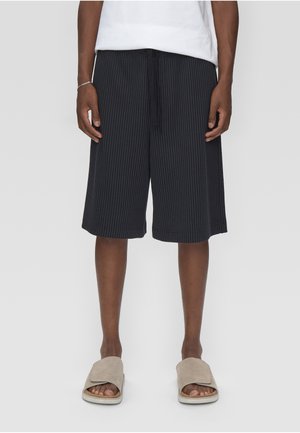 Man draagt zwarte knielange korte broek met krijtstreep, wit overhemd, beige slides sandalen, staand tegen een effen lichte achtergrond.