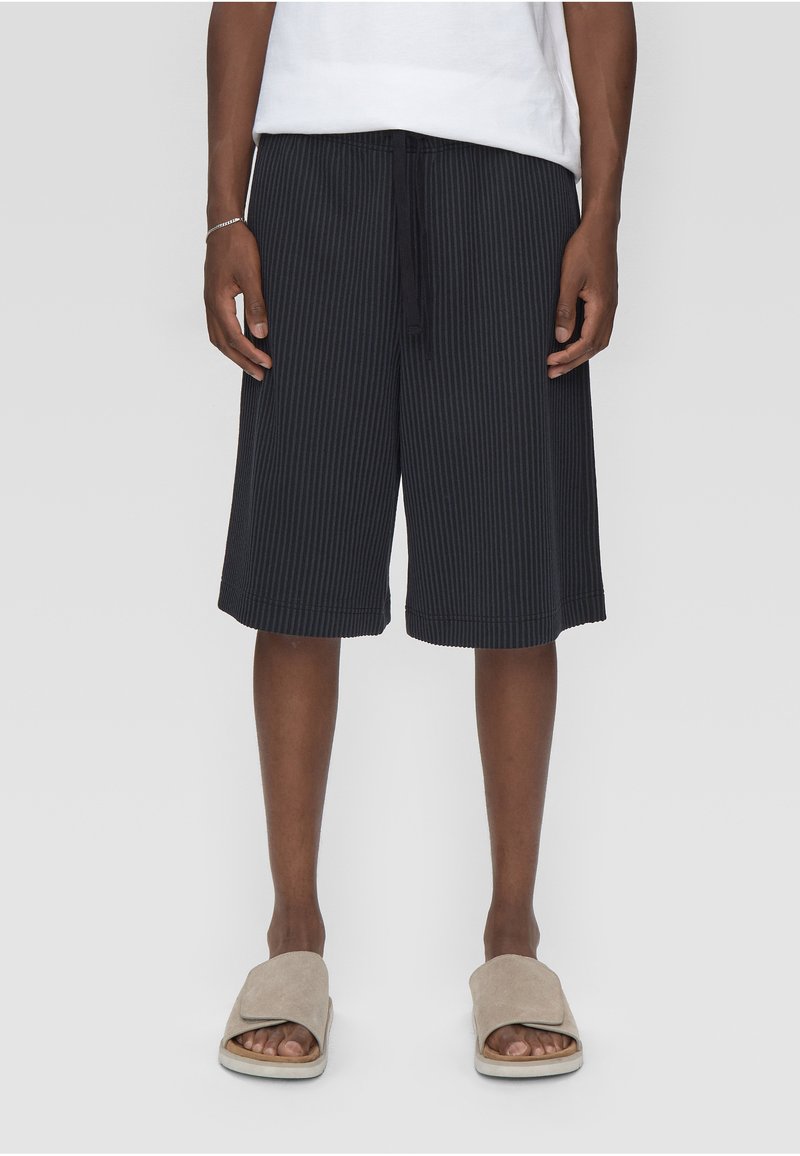 Homme portant un short noir à fines rayures jusqu'aux genoux, une chemise blanche, des sandales beige à enfiler, debout devant un fond clair uni.