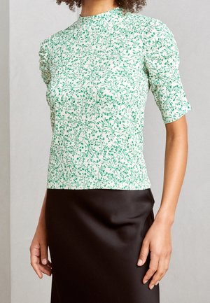 Vrouw die een witte blouse met korte mouwen draagt met een groene bloemenprint en opgeblazen schouders, gecombineerd met een zwarte rok.