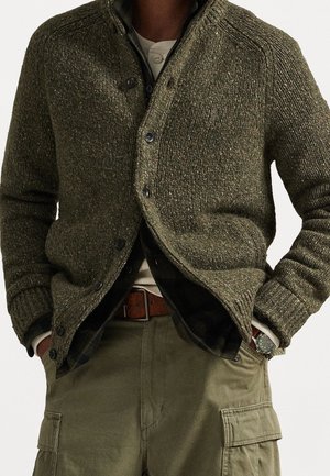 Person trägt einen strukturierten olivgrünen Cardigan über einem mehrlagigen Hemd und Pullover, mit khakifarbenen Cargohosen und Händen in den Taschen.