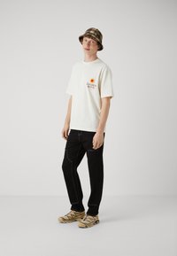 Hvit bomull t-skjorte med grafisk trykk og soldesign, sammen med svarte bukser med hvite sting, og beige sneakers med fargerike detaljer.