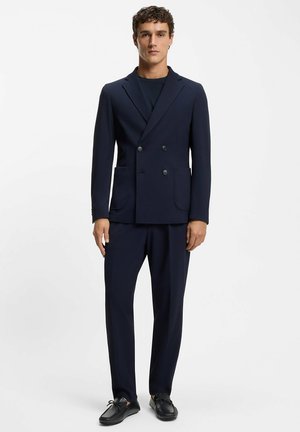 Man staat en draagt een marineblauwe dubbelgeknoopte blazer, bijpassende broek, zwarte loafers en een donkerblauw shirt tegen een witte achtergrond.