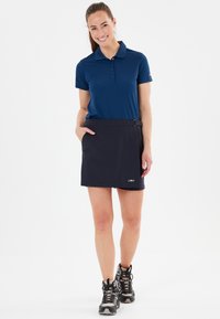 Mörkblå polo-shirt med knappar och korta ärmar, i kombination med en mörk kjol som har ett bälte och fickor. Modellen bär vandringskängor.
