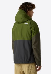 Giacca impermeabile verde e grigia con cappuccio, caratterizzata da blocchi di colore a contrasto e un logo sul retro. Tessuto liscio, stile casual.