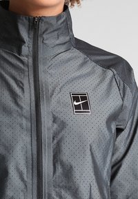 Chaqueta negra de Nike con cierre frontal, tela texturizada con perforaciones y un parche de logo en la parte superior izquierda.