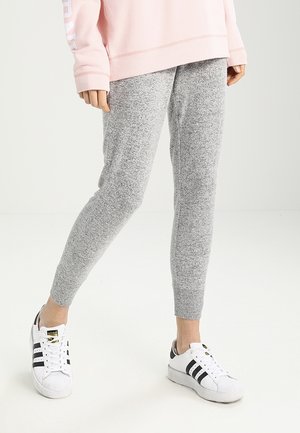 Personne portant un pantalon de jogging en maille gris, des baskets blanches Adidas avec des bandes noires, et un sweat rose clair avec un texte blanc sur la manche.